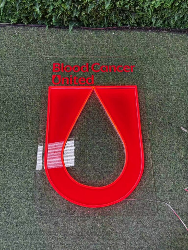Blood Cancer United charity sign displayed on grass, with UV-printed wording and red logo panels outlined by LED neon.